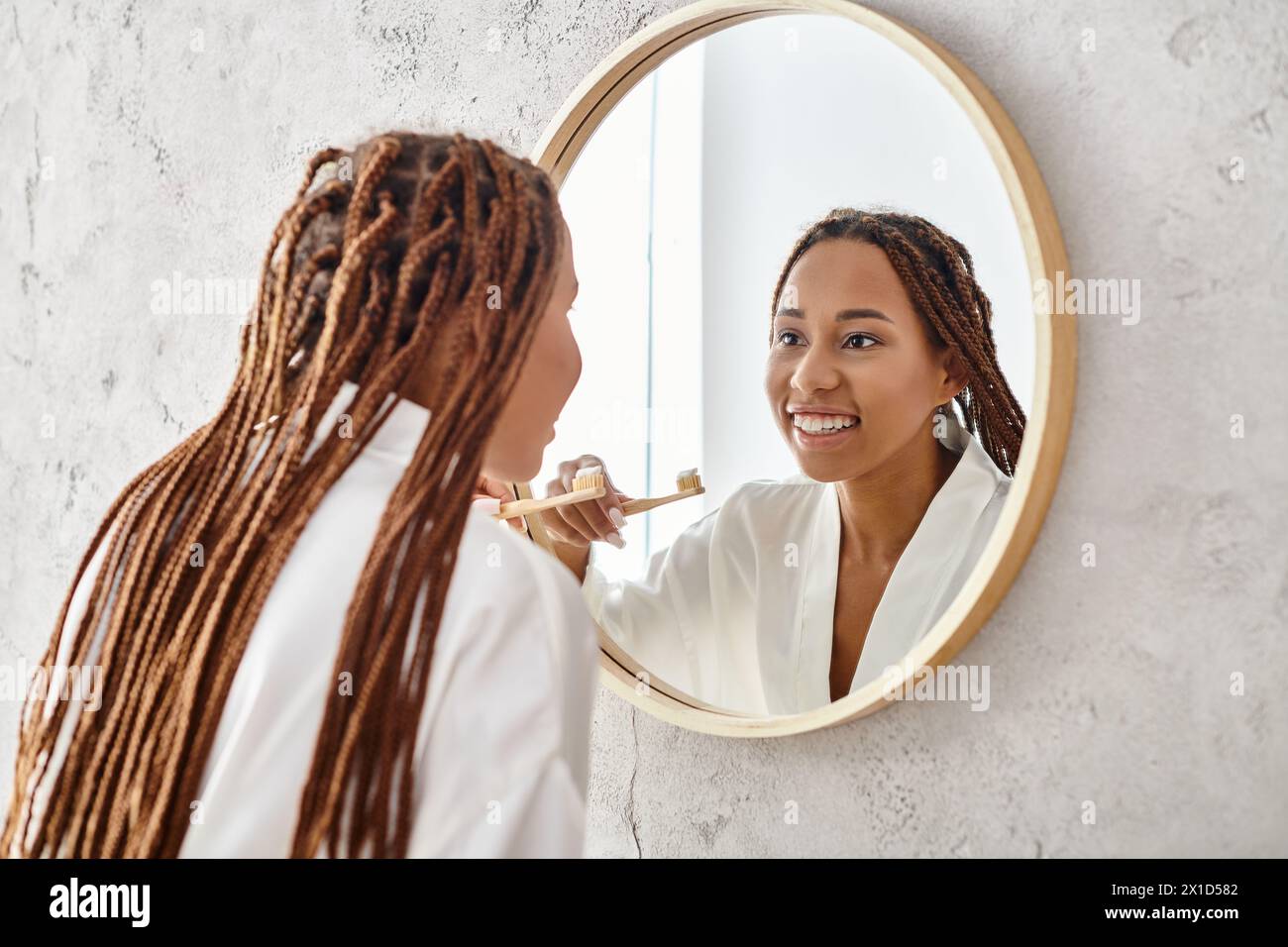 Una donna afroamericana con trecce afro in un accappatoio che si pulisce i denti in un moderno specchio del bagno. Foto Stock