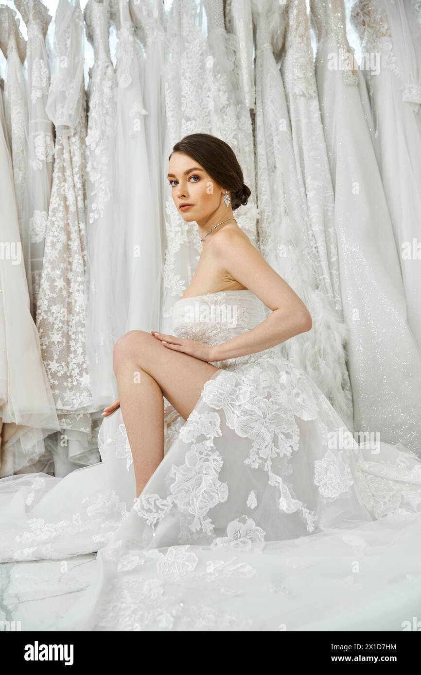 Una giovane sposa bruna in un abito da sposa bianco si siede graziosamente su un letto, contemplando l'occasione più importante che ci aspetta. Foto Stock