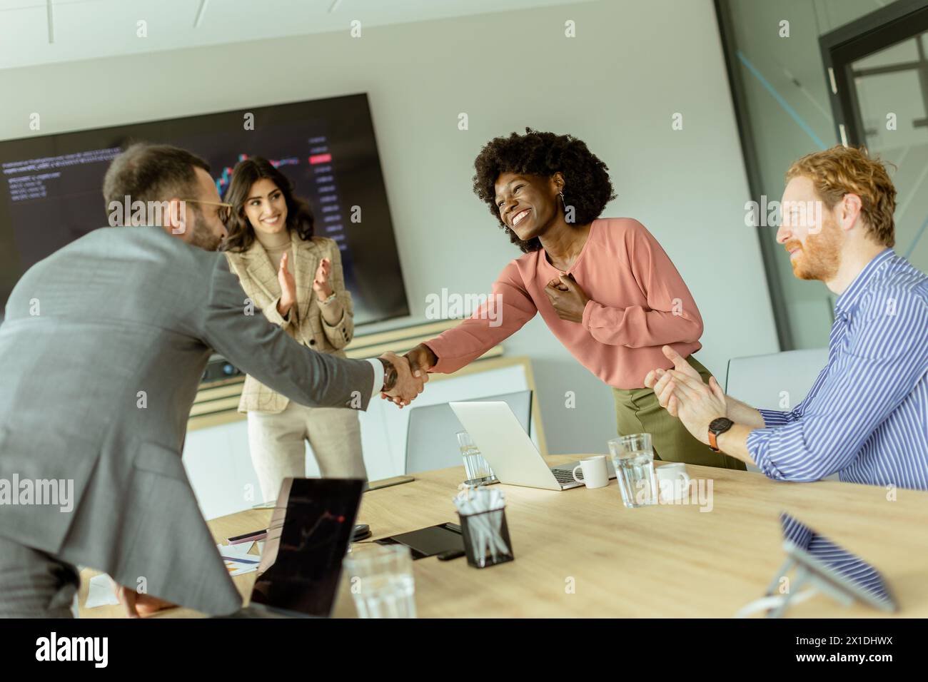 Due colleghi stringono la mano in accordo, mentre un altro paio applaude al loro successo in una sala conferenze ben illuminata Foto Stock