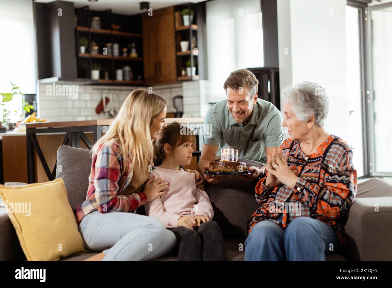 Una scena emozionante si svolge mentre una famiglia multigenerazionale si riunisce su un divano per presentare una torta di compleanno a una nonna adorata, creando ricordi indimenticabili Foto Stock
