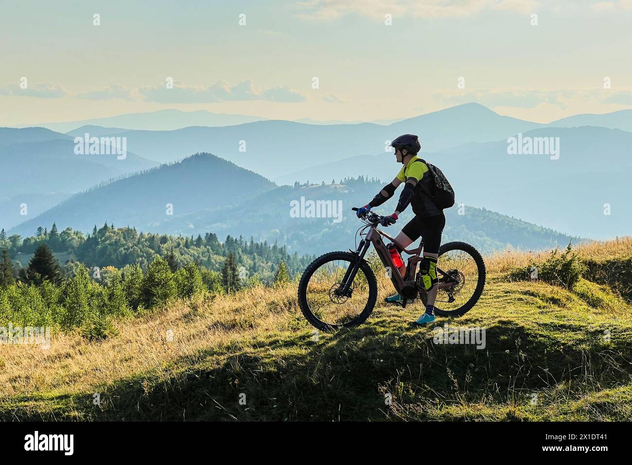 Ciclista che guida una bicicletta elettrica all'aperto nelle giornate di sole. Turista maschile che riposa su una collina erbosa, godendo di un bellissimo paesaggio montano, indossando casco e zaino. Concetto di svago attivo. Foto Stock
