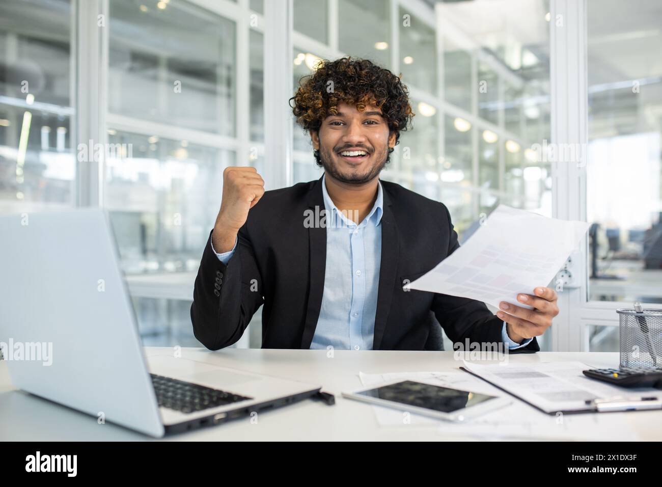 Un giovane allegro con una tuta da lavoro celebra un progetto o un risultato di successo seduto alla sua scrivania in un ambiente moderno, mostrando il pugno della vittoria e sorridendo con i documenti in mano. Foto Stock
