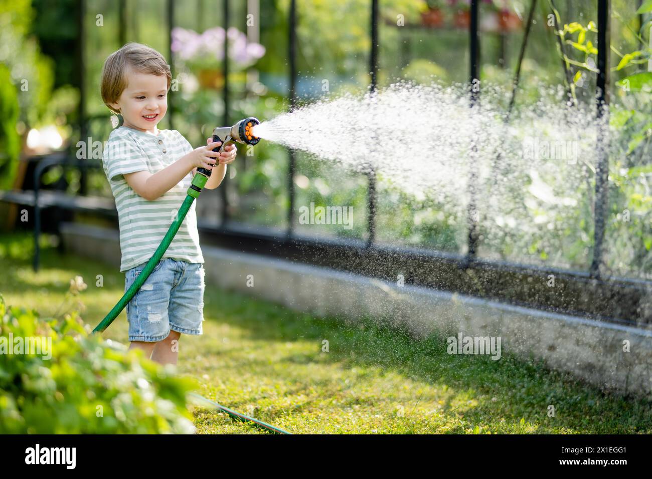 Un bambino adorabile che innaffiava aiuole di fiori in giardino durante il giorno d'estate. Bambino che usa il tubo da giardino per innaffiare le verdure. Ragazzo che aiuta con le faccende quotidiane. Mamma Foto Stock
