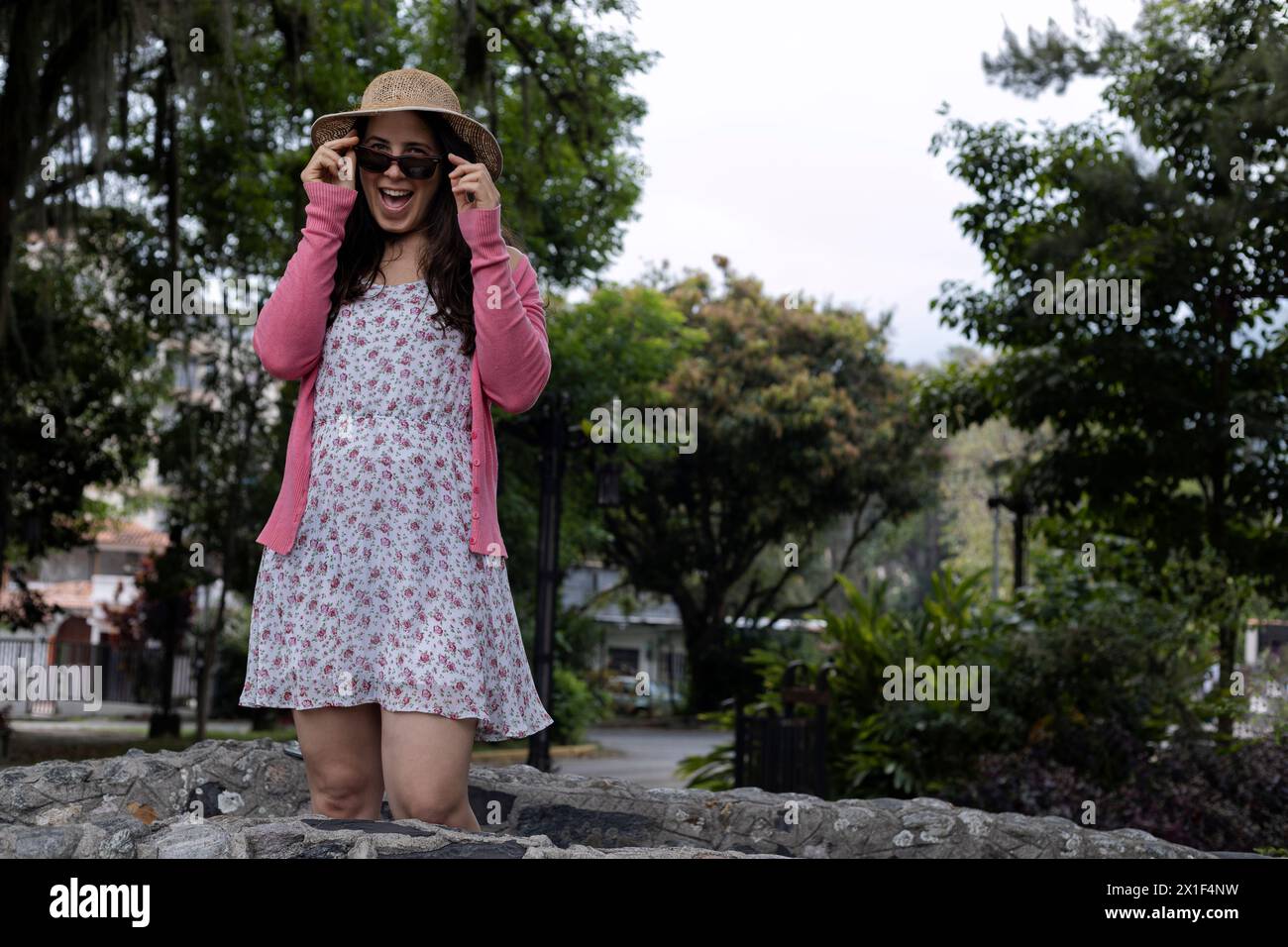 Giovane donna latinoamericana (33) sta con un atteggiamento positivo guardando i suoi occhiali. Indossa cappello, abito floreale e maglione rosa. Concetto primaverile. Foto Stock
