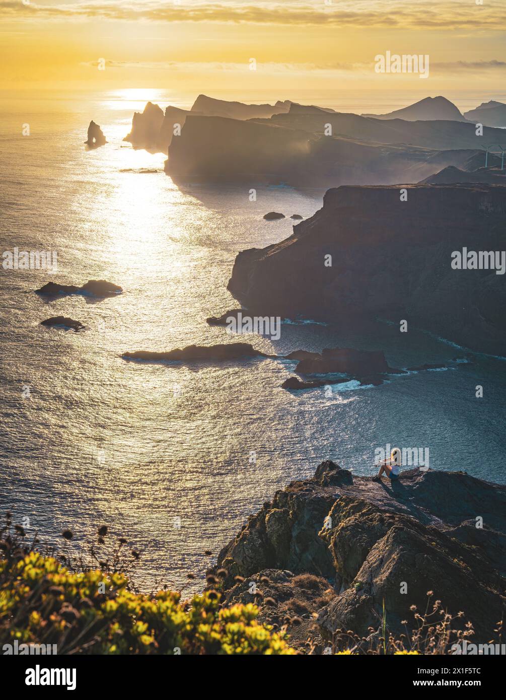 Descrizione: Le donne turistiche si trovano in un bellissimo punto panoramico su una scogliera ripida che si affaccia sul mare e sulle aspre colline pedemontane della costa di Madeira all'alba. Foto Stock