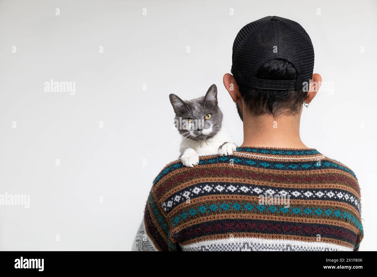 Uomo latinoamericano in piedi sulla schiena con il berretto che tiene il gatto mentre si appoggia sulla spalla guardando la macchina fotografica. Sfondo bianco. Concetto di animali domestici Foto Stock