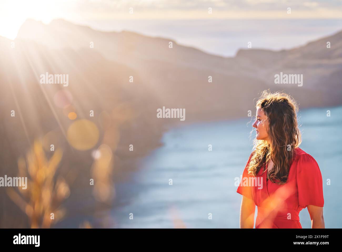 Descrizione: Vista laterale di una donna in abito rosso che si gode il bagno di sole mattutino su un'isola vulcanica nell'Oceano Atlantico. São Lourento, Isola di Madeira, po Foto Stock