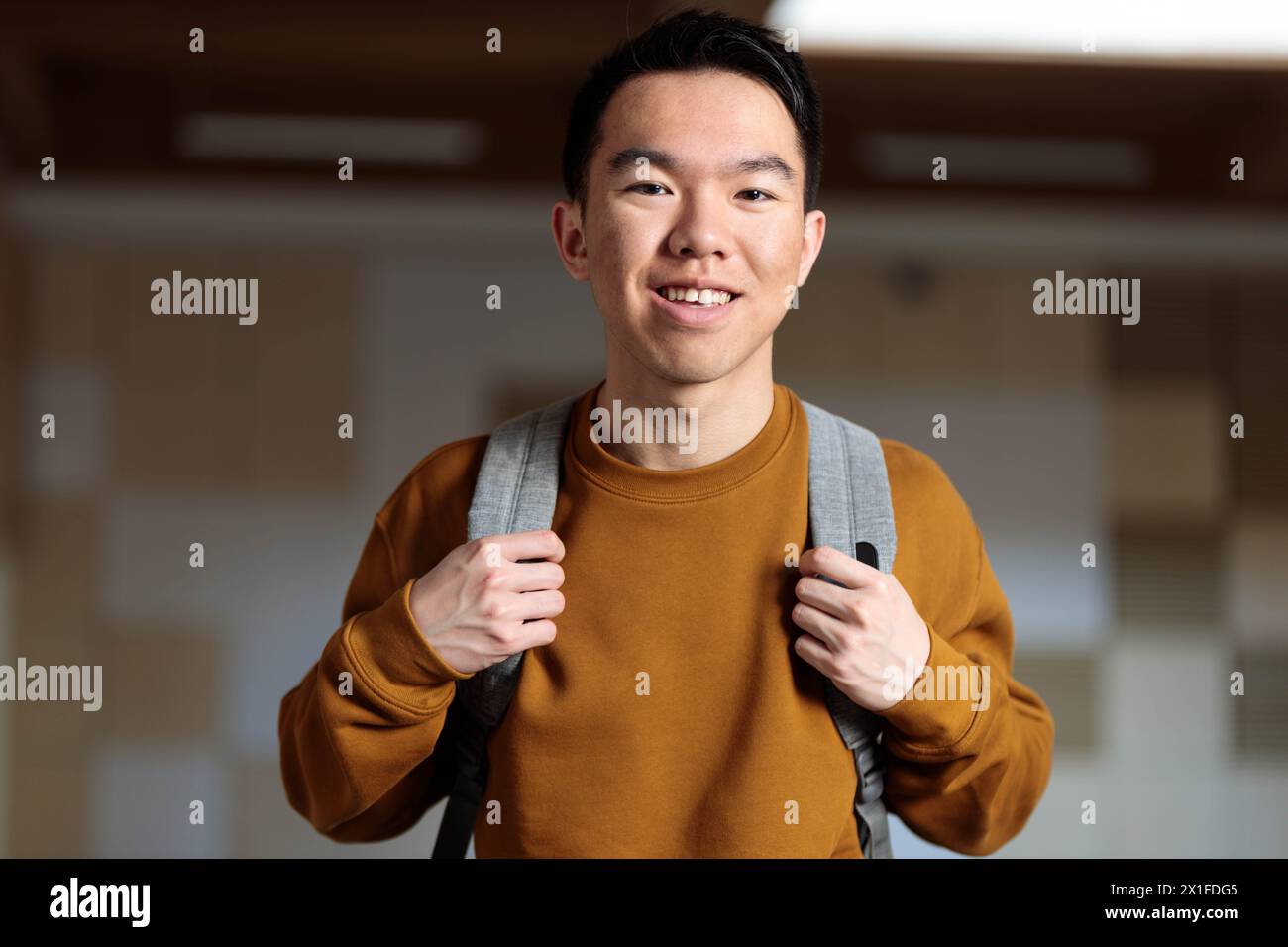 Una giovane studentessa asiatica in maglione marrone con zaino, è felice e pronta per la scuola guardando la macchina fotografica Foto Stock