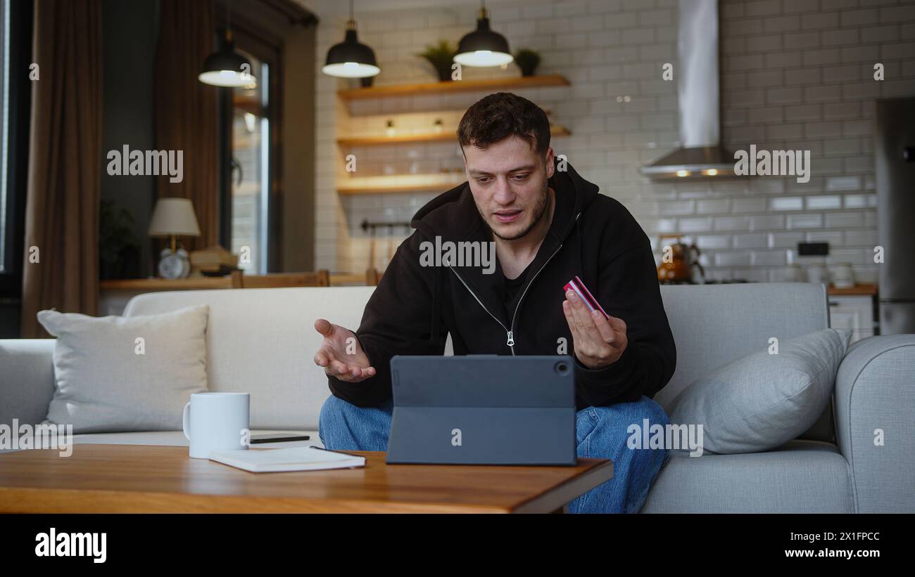 Un giovane uomo seduto sul divano di casa, inserendo il numero di carta di credito sul tablet per acquistare online la carta di credito viene rifiutata o fuori limite, sentendosi arrabbiato Foto Stock