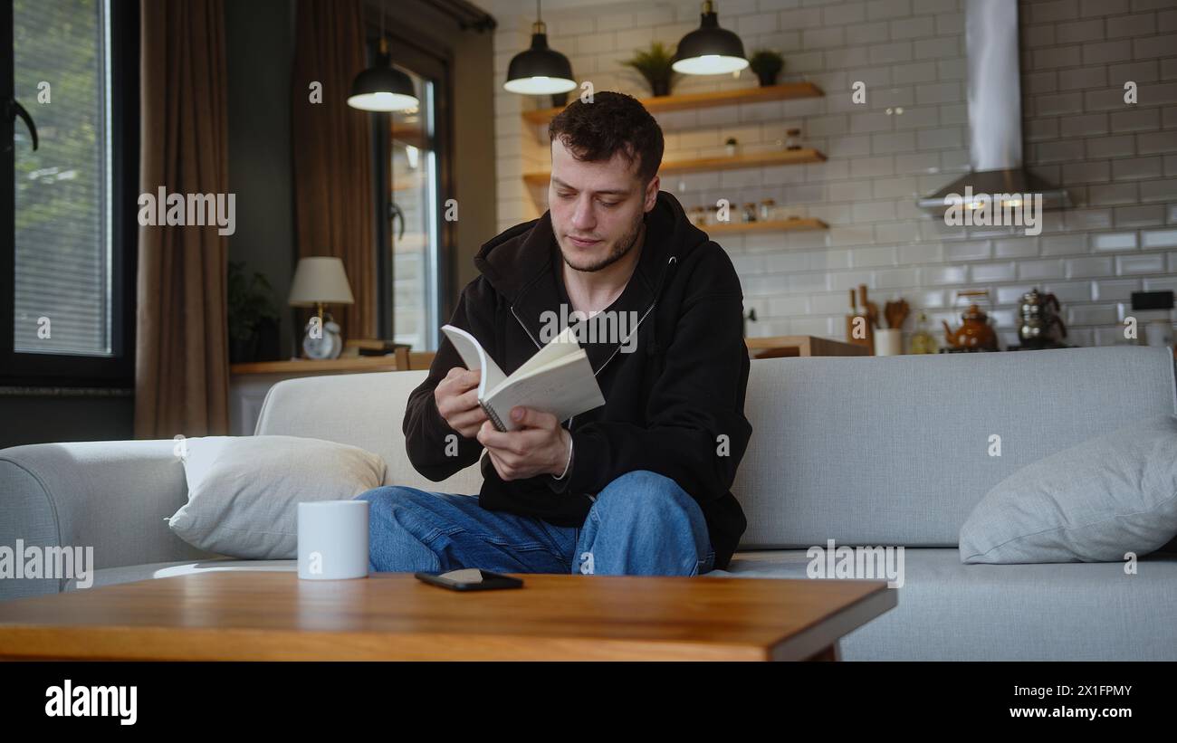 Il giovane seduto sul divano prende il notebook dal tavolo, guarda attraverso le pagine, strappa una pagina e getta il notebook sul tavolo a casa. Foto Stock