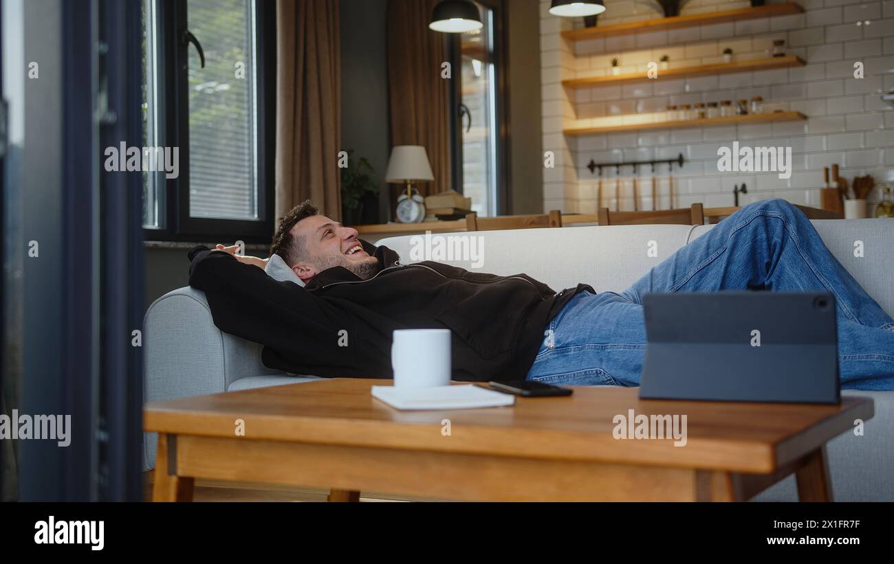 Un giovane uomo cade sdraiato su un comodo divano e riposante dopo una dura giornata di lavoro, fa una pausa nell'accogliente salotto di casa. Foto Stock