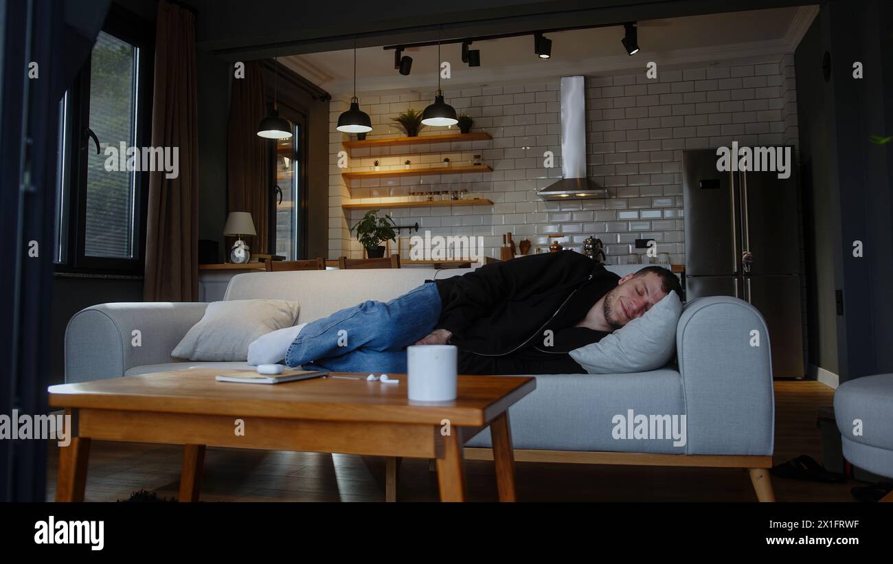 Un uomo assonnato seduto sul divano sbadigliato e sdraiato a fare un pisolino con abiti casual. Riposati e rilassati dopo una dura giornata di lavoro facendo una pausa in un salotto Foto Stock