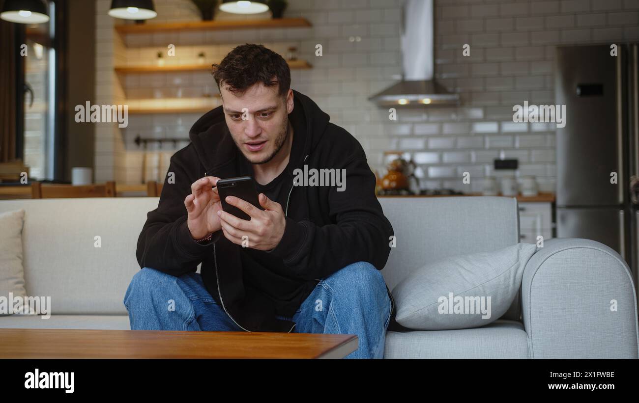Il giovane seduto sul divano reagisce a ciò che vede, navigando su Internet sul telefono cellulare, controllando le e-mail, leggendo notizie sui media, scorrendo i social media, usando Mo Foto Stock