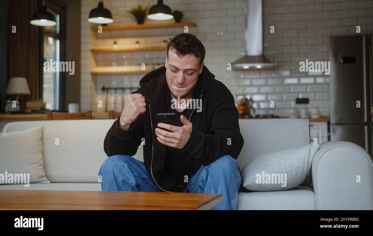 Uomo seduto sul divano che usa lo smartphone controllare messaggio leggere avviso ricevere buone notizie via e-mail celebrare grandi opportunità si sente felice Foto Stock