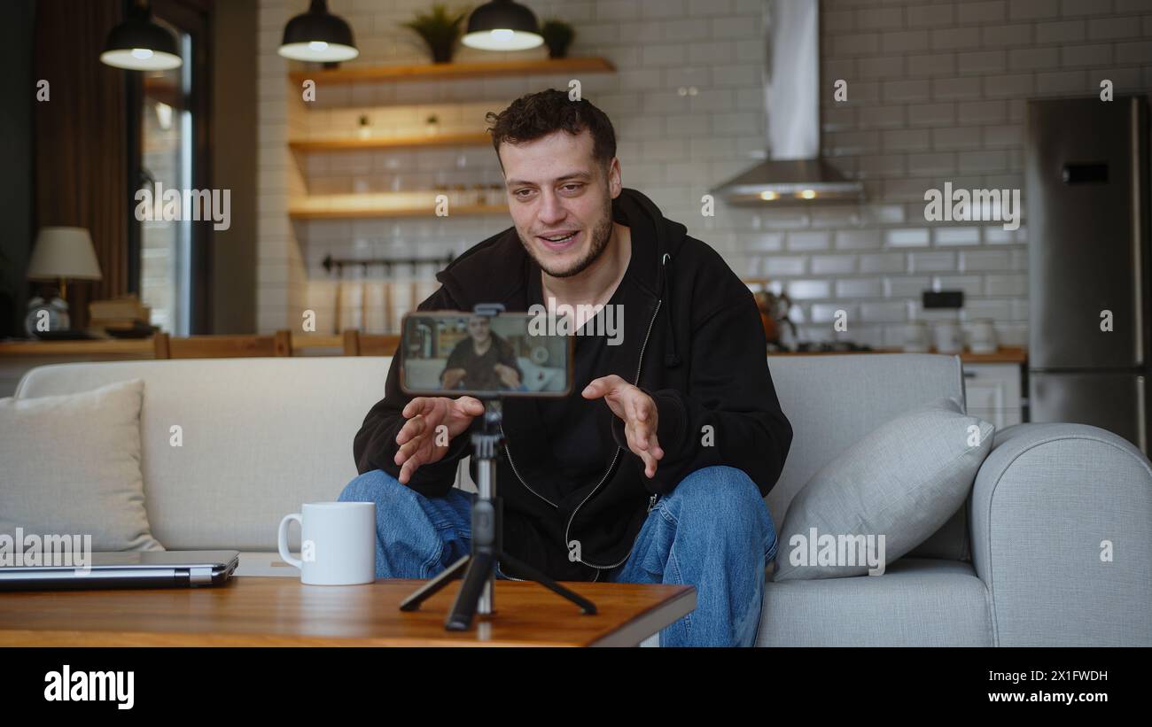 Il giovane vlogger influencer si siede sul divano registrando video blog sul treppiede della fotocamera mobile parla per il canale multimediale personale, in streaming live session blo Foto Stock