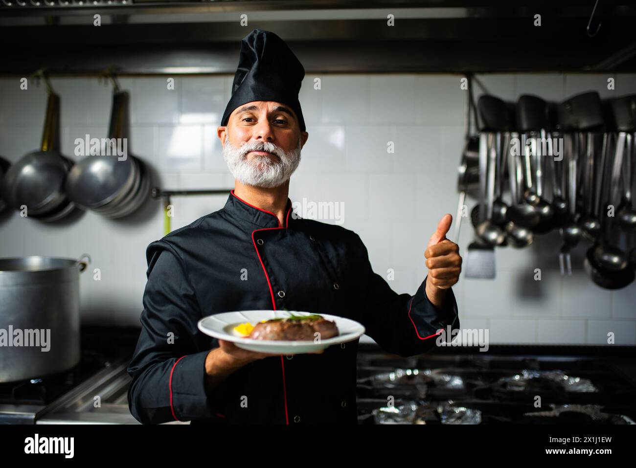 Lo chef professionista in uniforme regala il pollice mentre tiene un pasto servito nella cucina di un ristorante Foto Stock