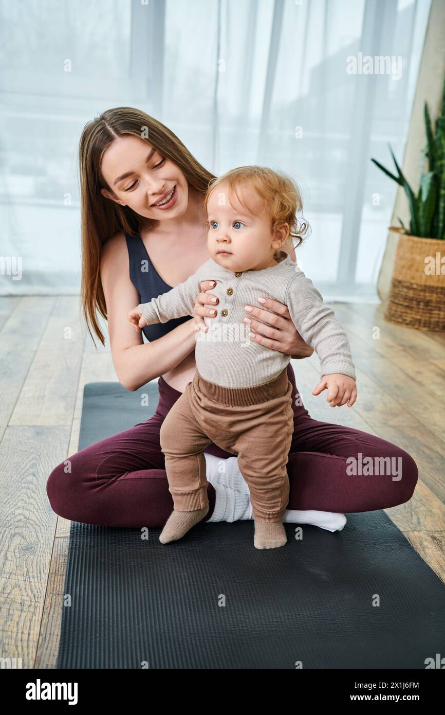 Una giovane madre tiene delicatamente il bambino su un tappetino da yoga, guidato da un coach ai corsi dei genitori nel comfort della loro casa. Foto Stock