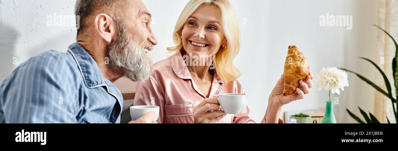 Una coppia matura e amorevole vestita con intimi casalinghi condivide un pasto insieme al loro tavolo da cucina, godendosi un momento di Unione. Foto Stock