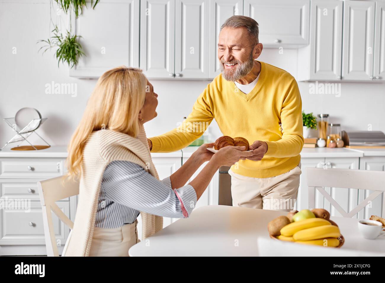 Un uomo e una donna maturi in un abbigliamento accogliente si trovano a un tavolo da cucina, condividendo un momento tenero mentre preparano un pasto insieme a casa. Foto Stock