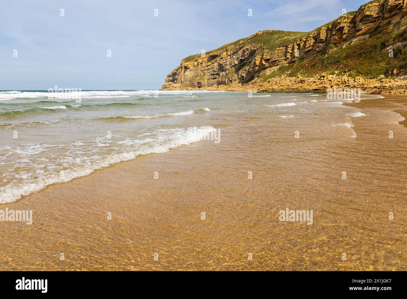 Spiaggia di Luaña con sabbia dorata, acqua scintillante alla luce del sole, onde e scogliere. Comune di Alfoz de Lloredo, Cantabria, Spagna. Foto Stock