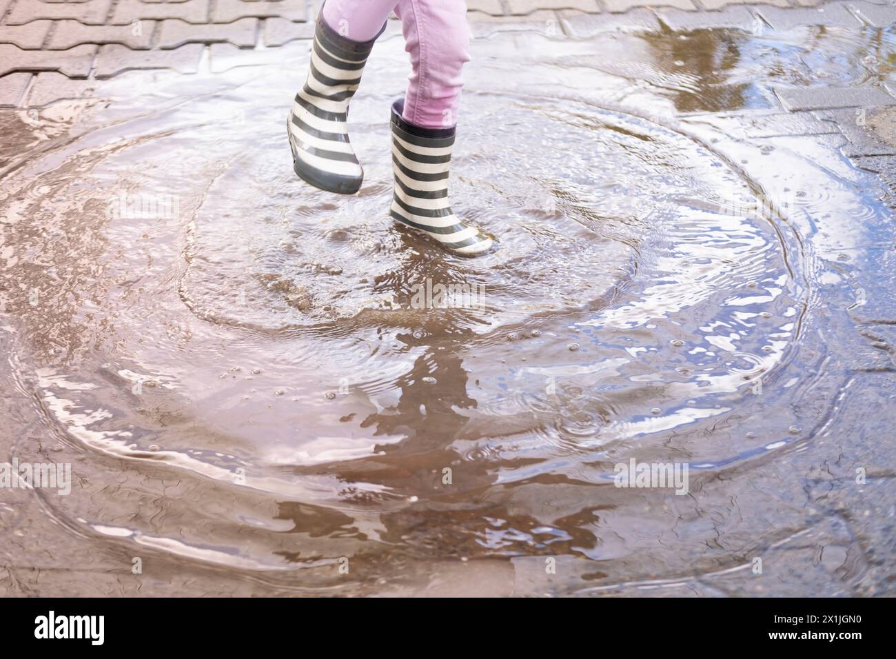 primo piano, la bambina di 5 anni salta gioiosamente in un pozzetto indossando stivali di gomma, i piedi dei bambini in schizzi d'acqua, saltando felicemente, i piaceri dell'infanzia Foto Stock