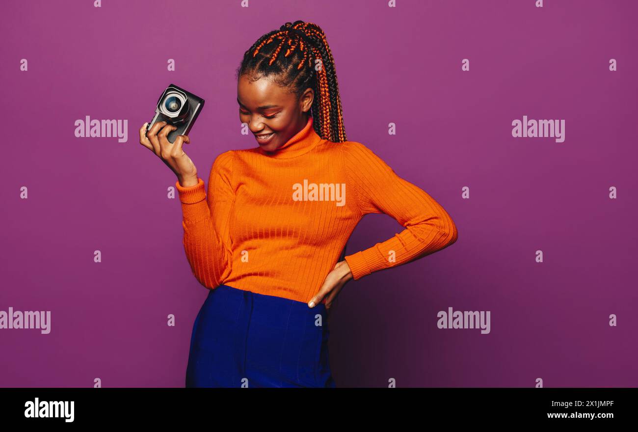 Ragazza africana con colorate trecce bicolore, in uno studio di sfondo viola. Tenere in mano una fotocamera digitale, scattare una foto di qualcosa fuori dalla fotocamera. Foto Stock