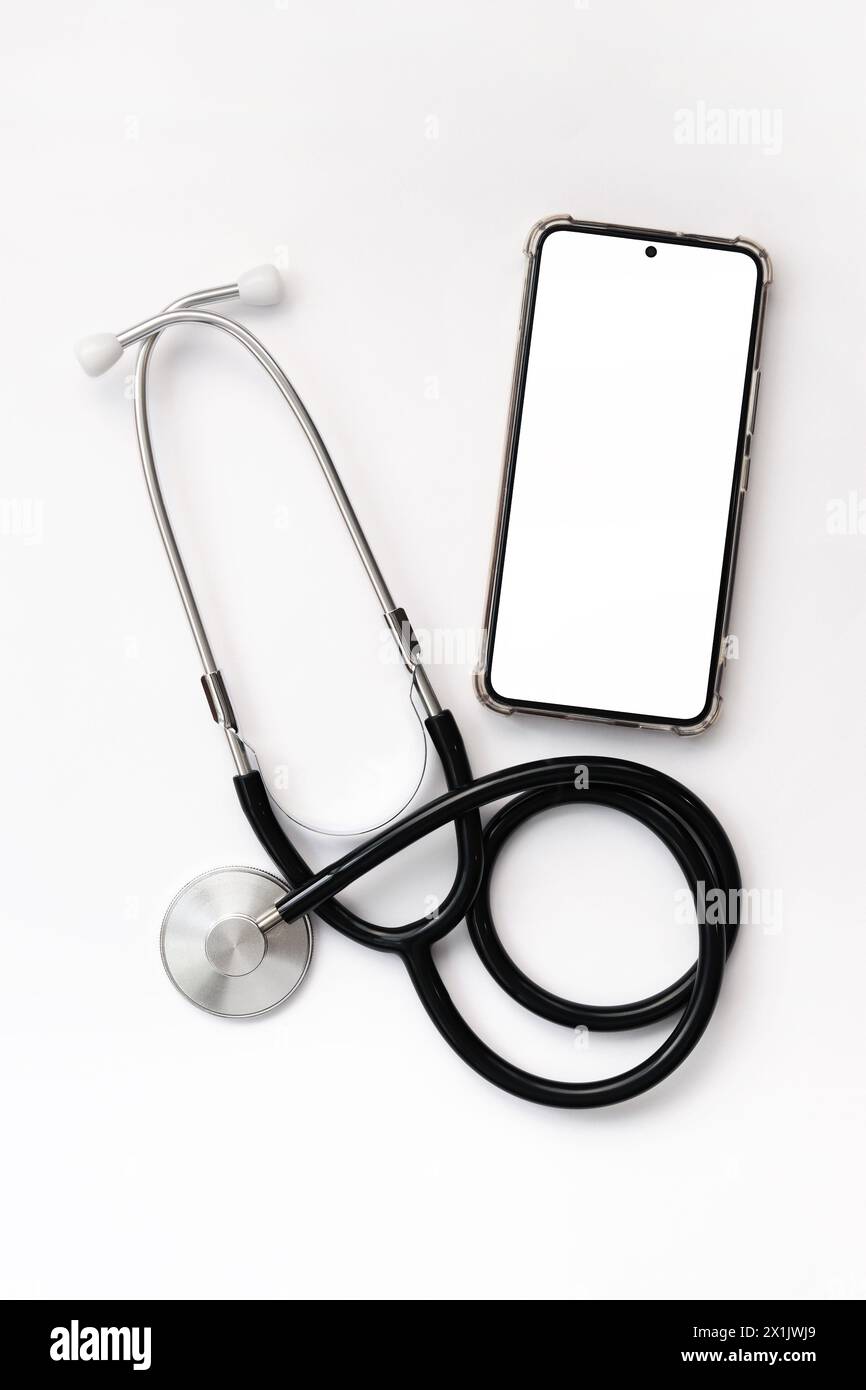 Stetoscopio su sfondo bianco e smartphone con schermo bianco, vista dall'alto. Strumento medico. Concetto di cardiologia e medicina, assistenza sanitaria. Foto Stock