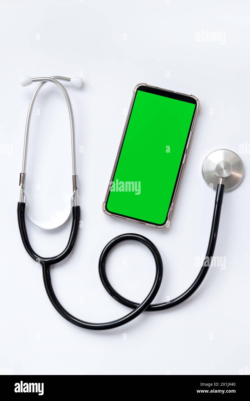 Stetoscopio su sfondo bianco e smartphone con schermo verde, vista dall'alto. Strumento medico. Concetto di cardiologia e medicina, assistenza sanitaria. AUS Foto Stock