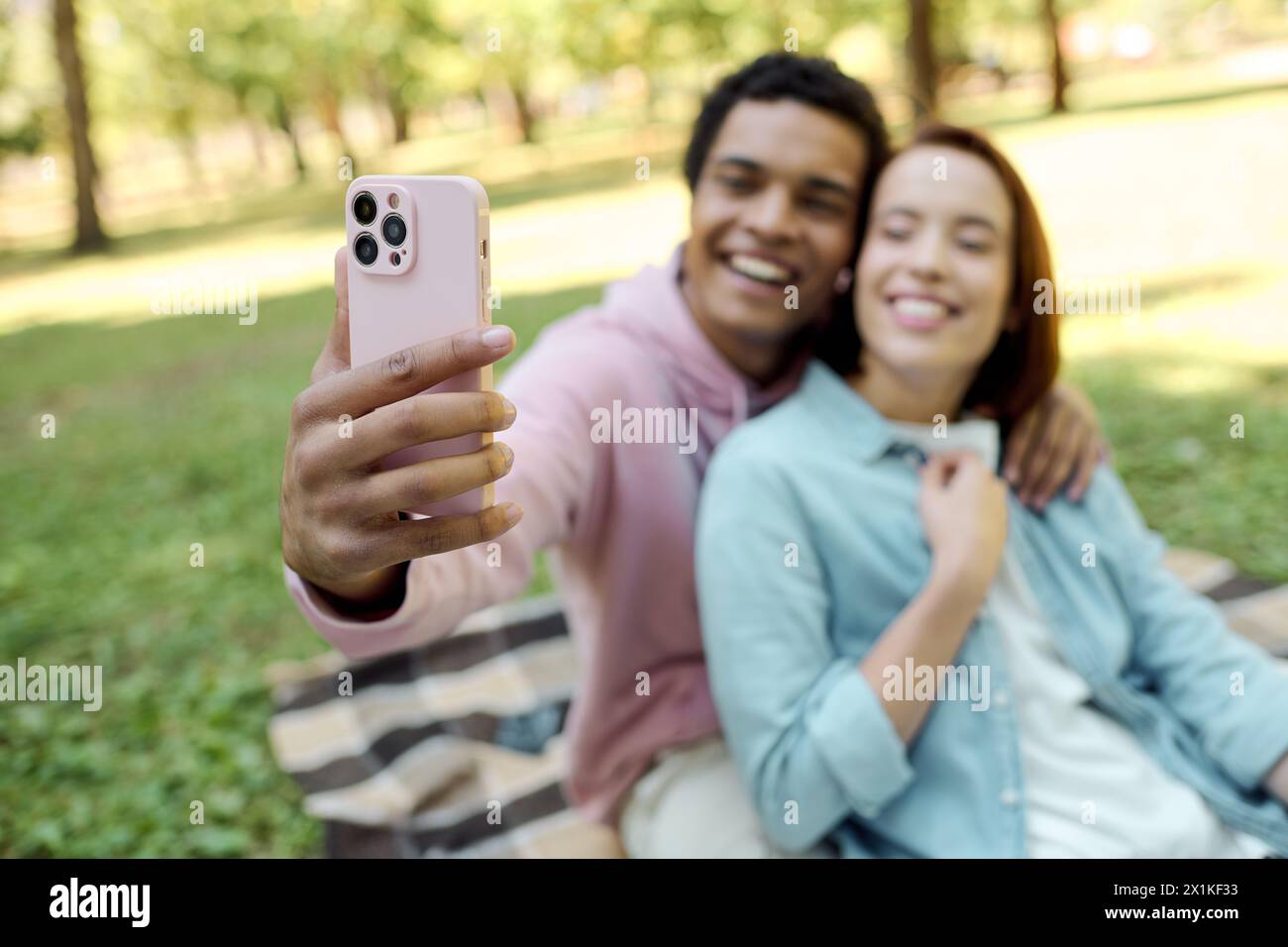 Una coppia eterogenea e amorevole con un abbigliamento vivace cattura un momento di felicità insieme scattando un selfie in un bellissimo parco. Foto Stock