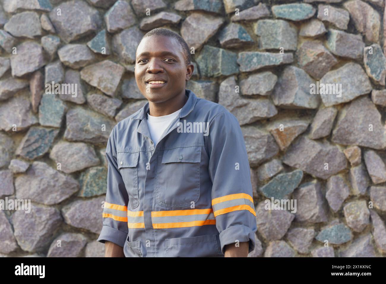 giovane africano con un grande sorriso e abbigliamento da lavoro, sfondo in pietra da parete Foto Stock