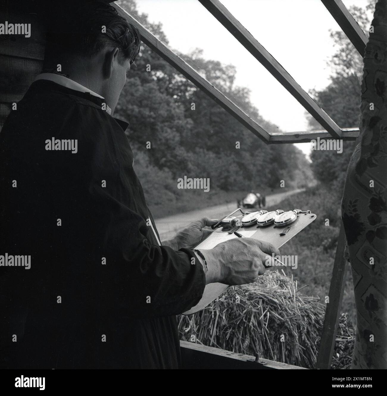 1962, storico, istruttore maschile di sport motoristici con tabellone e cronometri in cabina presso la pista della scuola guida di Geoff Clarke presso l'aerodromo di Finmere, Finmere, Bucks, Inghilterra, Regno Unito, cronometrare un'auto da corsa che fa un giro. Foto Stock