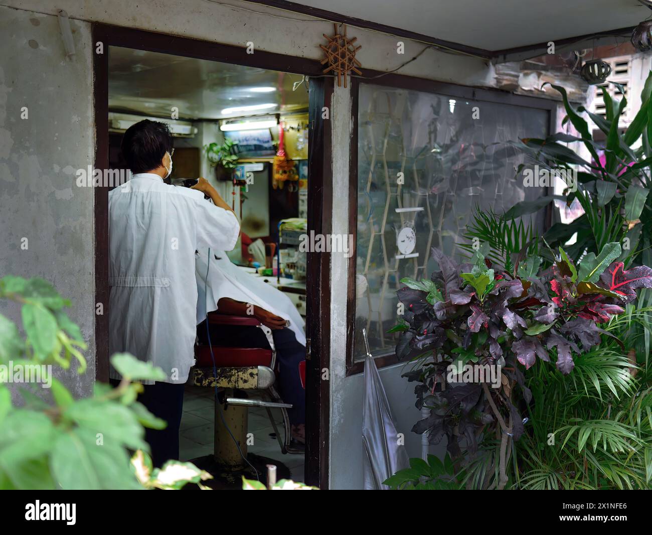 Un piccolo barbiere in vecchio stile nascosto nell'area locale di Bangkok, parrucchiere che indossa la maschera tagliando i capelli per i clienti, affari locali Foto Stock