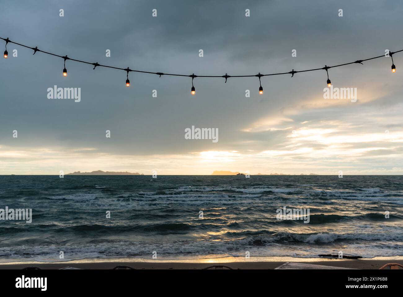 Affacciate su un mare tumultuoso, le luci a corda incorniciano la vista del tramonto, offrendo un mix di dramma della natura e atmosfera accogliente. Foto Stock