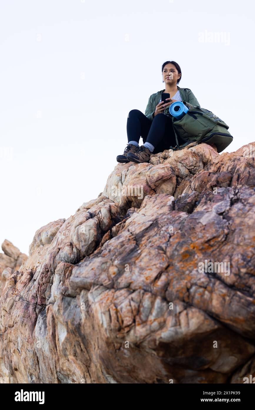Escursionista birazziale seduto su un picco roccioso in mezzo alla natura, tenendo in mano lo smartphone, spazio fotocopie Foto Stock