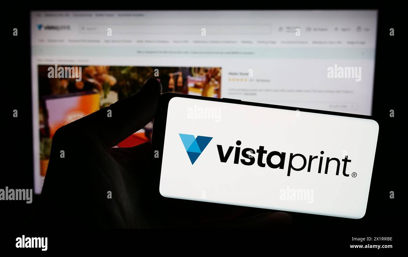 Persona che possiede uno smartphone con il logo della società di personalizzazione di massa Vistaprint (Vista) davanti al sito Web. Mettere a fuoco il display del telefono. Foto Stock