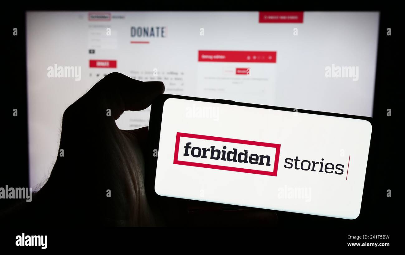 Persona che possiede un telefono cellulare con il logo dell'organizzazione di giornalismo investigativo Forbidden Stories davanti alla pagina web. Mettere a fuoco il display del telefono. Foto Stock