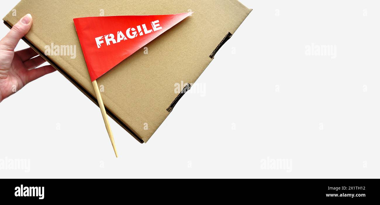 Una scatola di cartone in mano su sfondo monocromatico. Piccola bandiera di carta rossa con l'avvertenza "fragile" come etichetta, adesivo Foto Stock