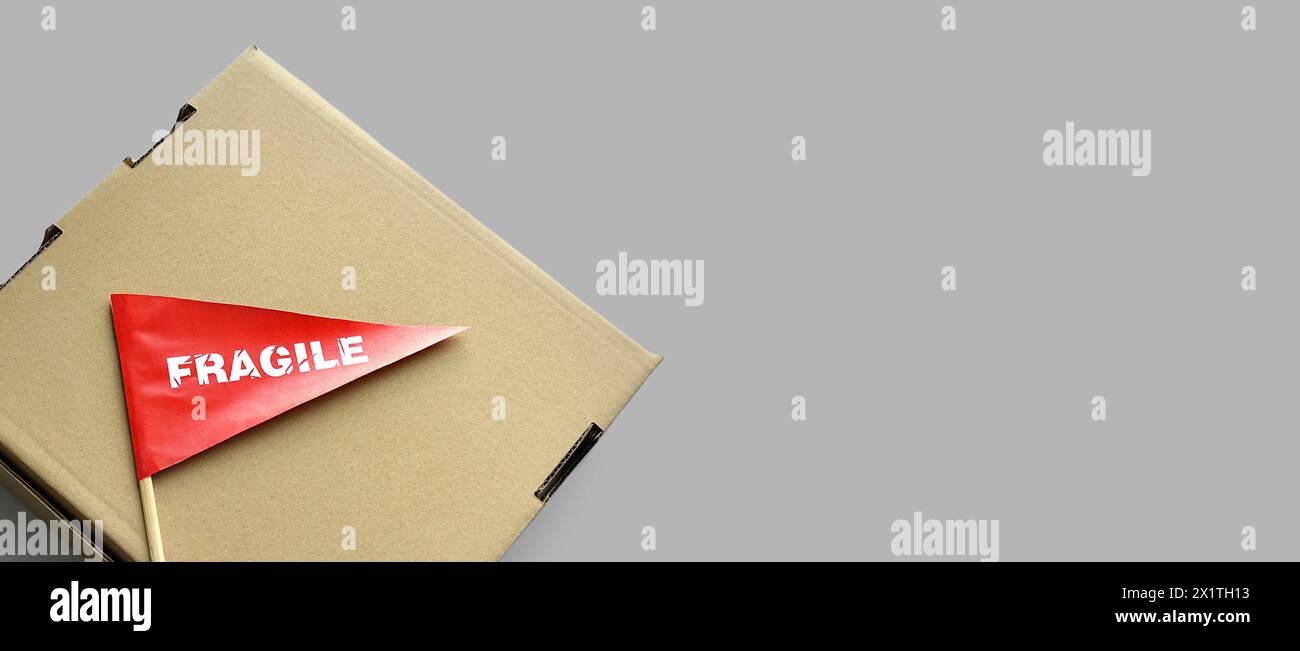 Una scatola di cartone su sfondo monocromatico. Piccola bandiera di carta rossa con l'avvertenza "fragile" come etichetta, adesivo Foto Stock