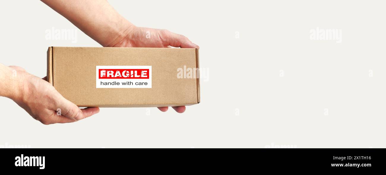 Una scatola di cartone rettangolare in mano per l'imballaggio di articoli fragili su uno sfondo chiaro e semplice. Etichetta di avvertenza rossa, adesivo per imballaggio "fragile" Foto Stock