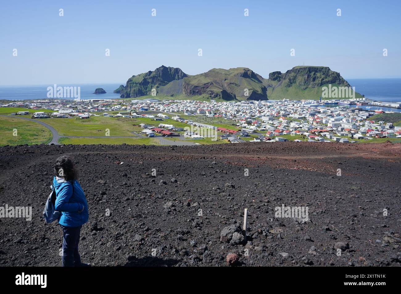 Una giovane donna guarda la città di Heimaey e la montagna di Heimaklettur dalla cima del cono vulcanico di Eldfell, le isole Westman, Islanda Foto Stock