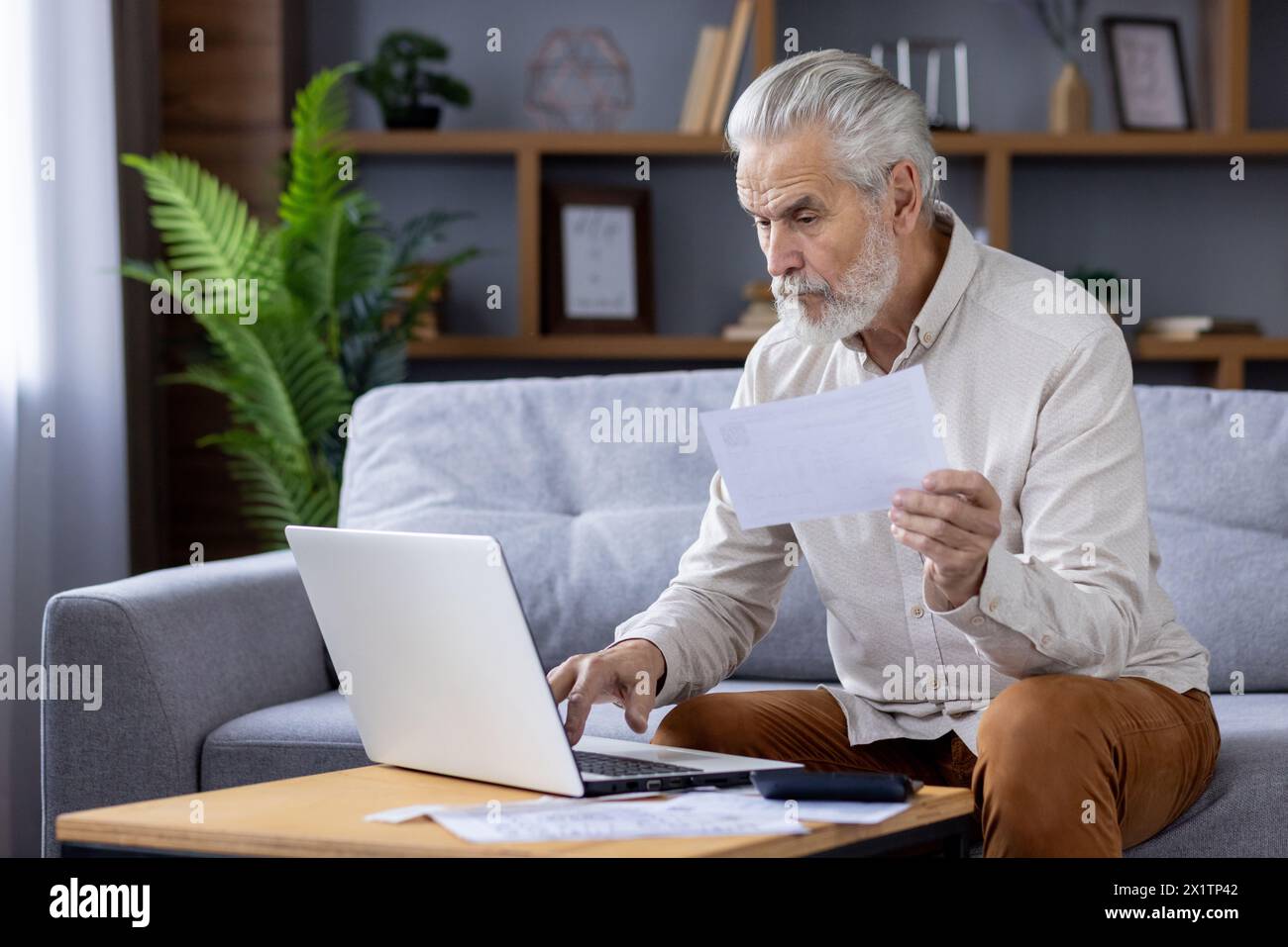 Uomo anziano concentrato con capelli grigi, che lavora su un notebook seduto su un divano, che esamina attentamente i documenti in un ambiente accogliente. Foto Stock