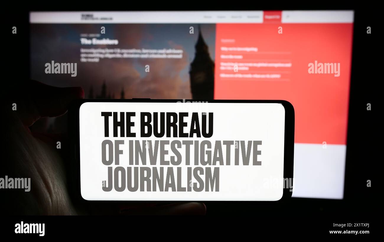 Persona che detiene un cellulare con il logo dell'organizzazione il Bureau of Investigative Journalism (TBIJ) di fronte alla pagina web. Mettere a fuoco il display del telefono. Foto Stock
