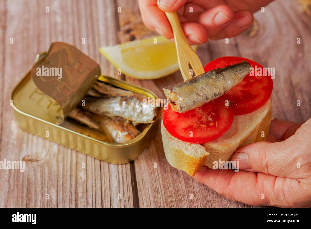 Una donna mette una sardina su una fetta di pane con pomodoro, sullo sfondo si può vedere la lattina di sardine e un limone su un tavolo di legno Foto Stock