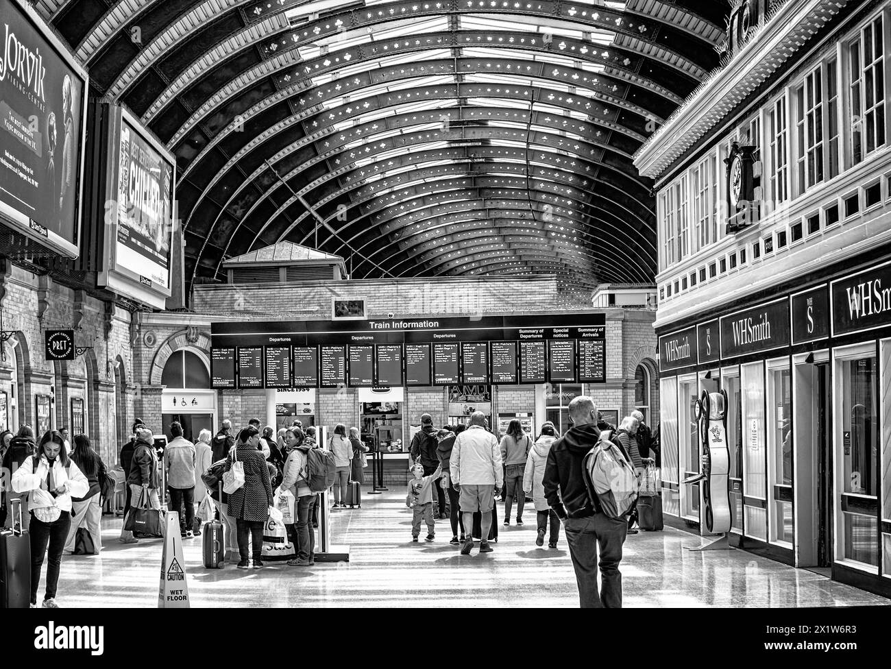 Un baldacchino di ferro copre un atrio della stazione ferroviaria con un cartello elettronico che mostra le informazioni sui treni. Un negozio è su un lato e una folla di persone Foto Stock