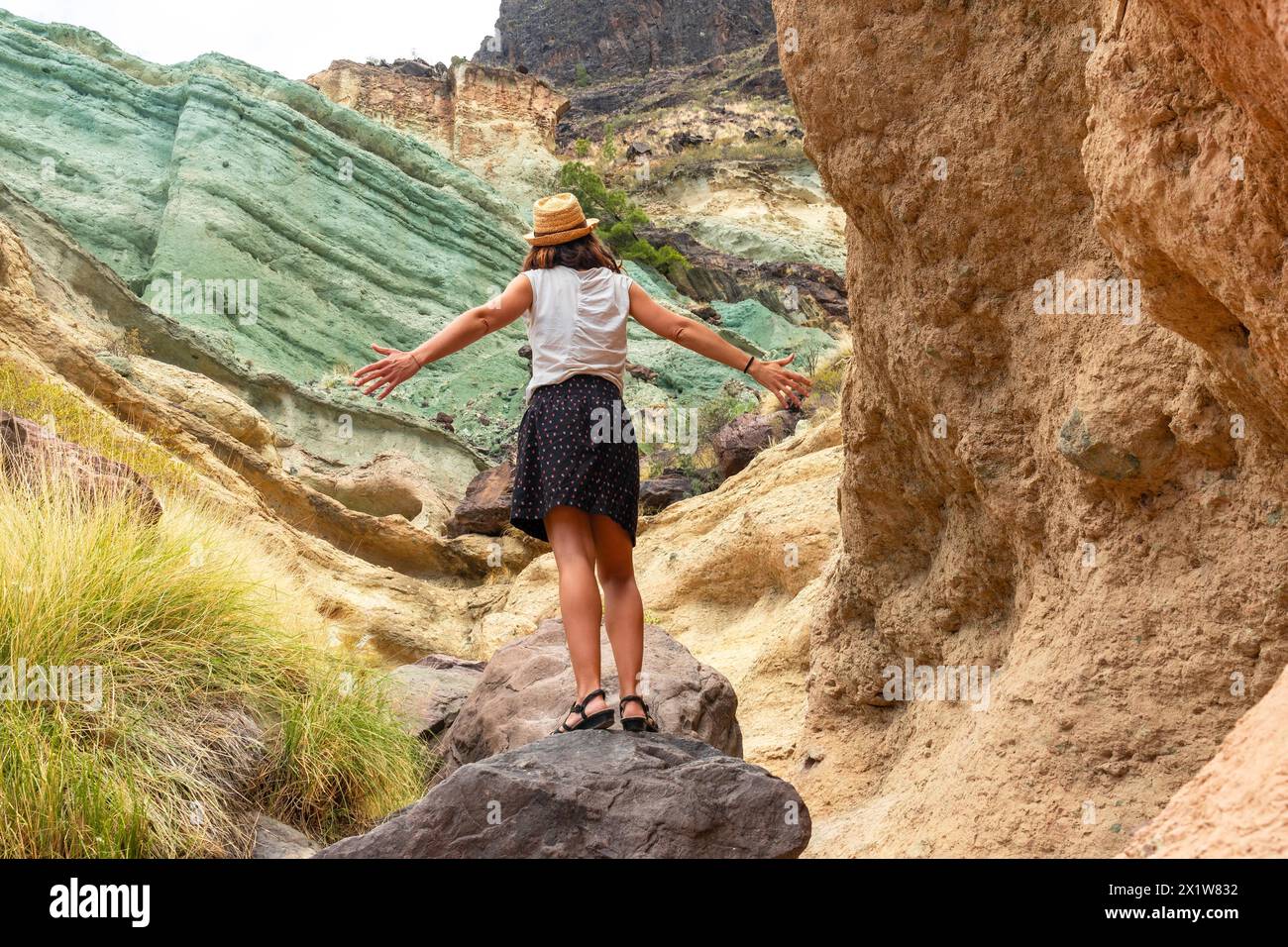 Una donna turistica con un cappello e braccia aperte all'Azulejos de Veneguera o Rainbow Rocks a Mogan, Gran Canaria Foto Stock