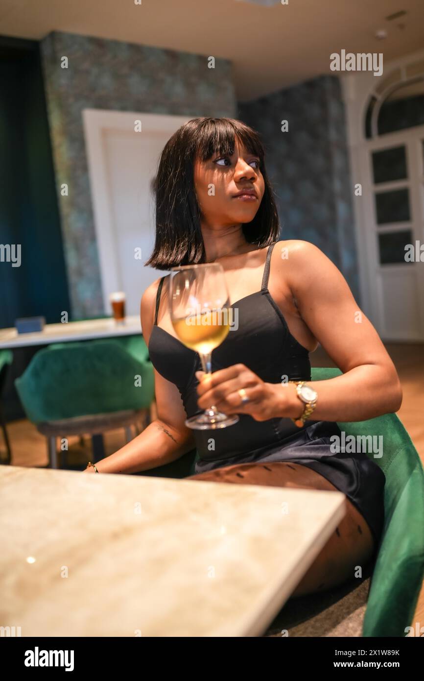 Foto verticale di una sensuale donna africana distratta con un bicchiere di vino bianco in un bar Foto Stock