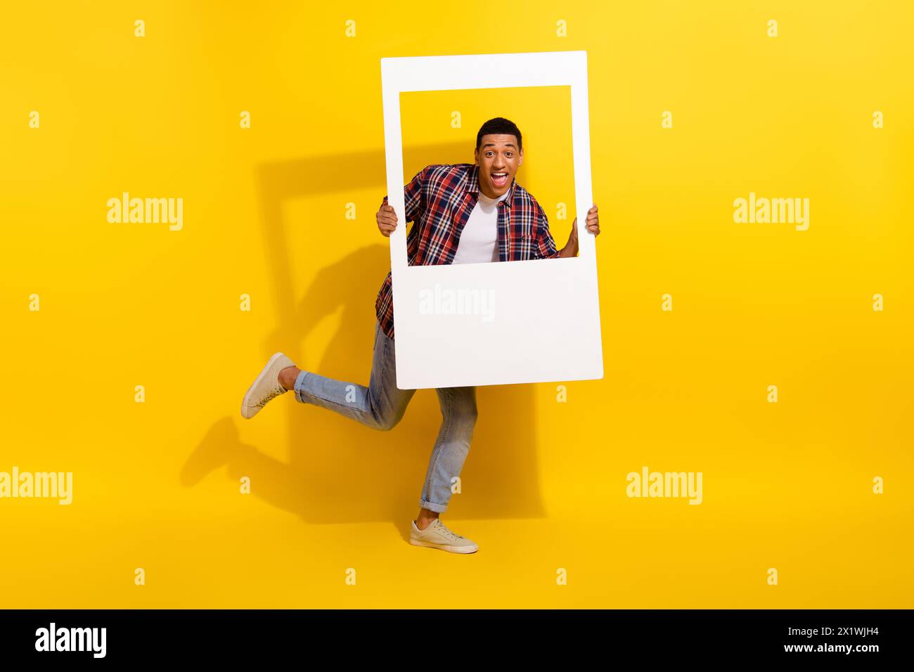 Foto a grandezza naturale di un eccentrico e allegro jeans con camicia a quadri dall'uomo che reggono la cornice bianca zona fotografica isolata su sfondo giallo Foto Stock