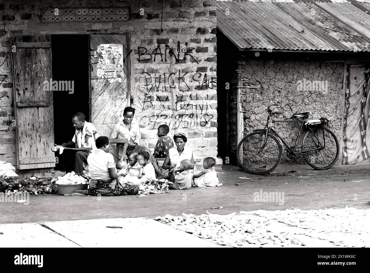 Una famiglia ugandese si riunisce fuori dalla loro umile casa, preparando verdure in vendita. Una bicicletta si appoggia contro il muro, pronta a trasportare merci sul mercato Foto Stock