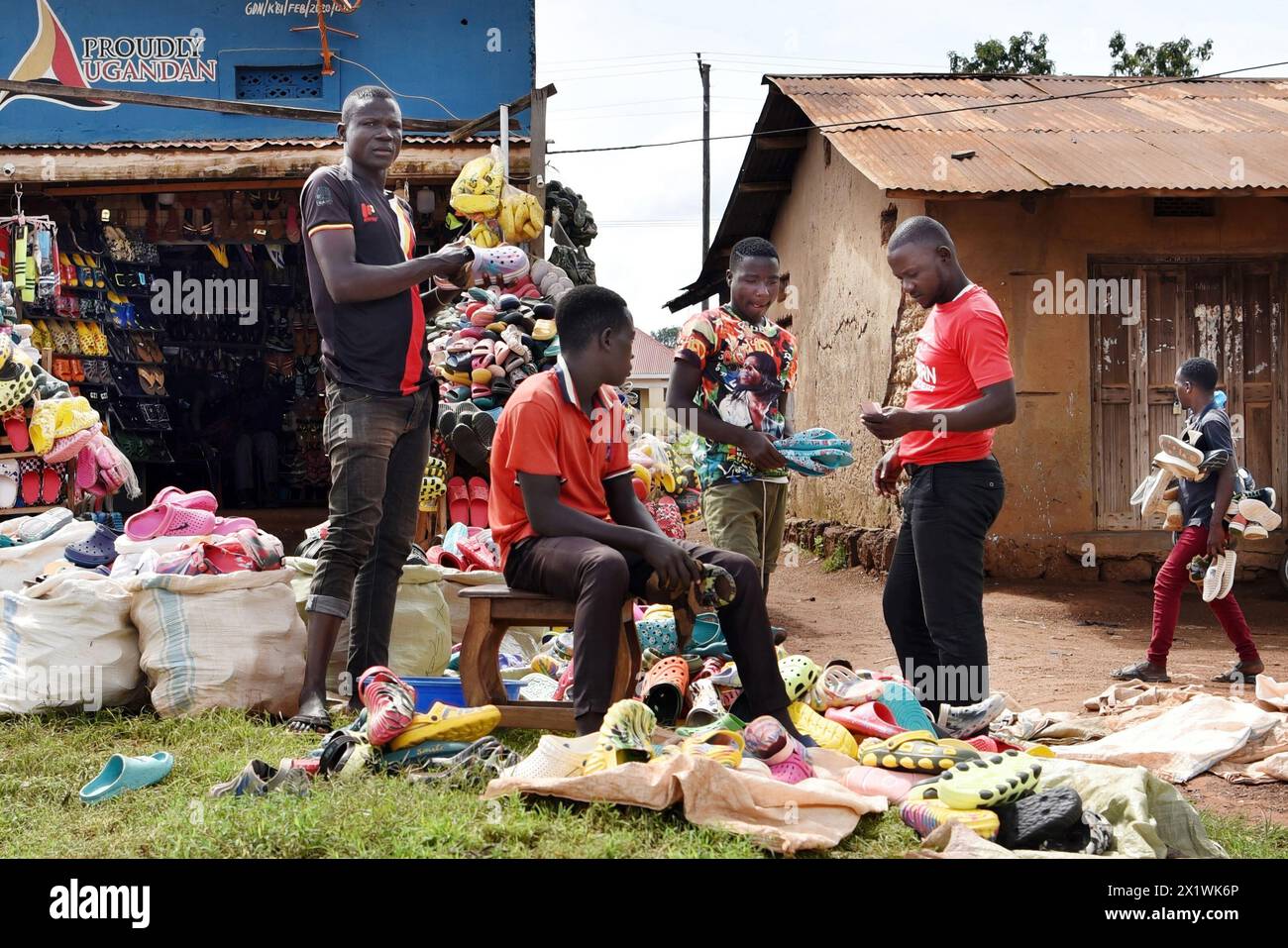 Una semplice capanna di fango si erge accanto a un umile negozio di scarpe da strada ugandese. Tre giovani vendono scarpe da ginnastica da grandi sacchi bianchi nelle vicinanze. Foto Stock
