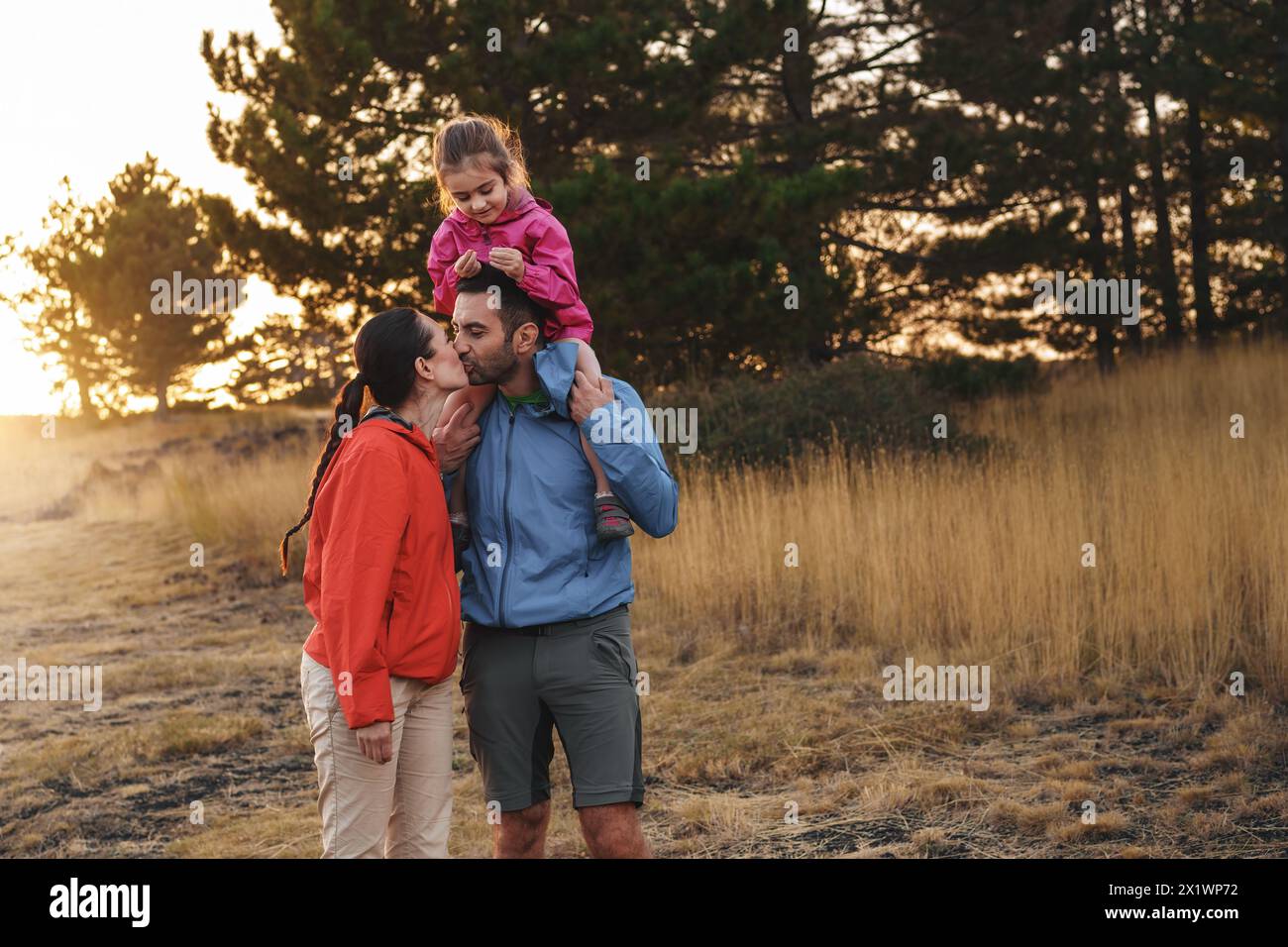 La famiglia ama una tranquilla passeggiata nella natura - bambino che cavalca felicemente sulle spalle di suo padre - la madre lo bacia: Affetto e solidarietà al tramonto. Foto Stock